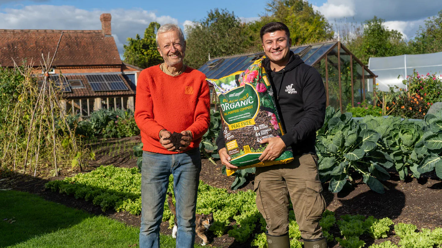Charles Dowding and Ashley Day pose in a garden, with Ashley holding a bag of Growmoor Organic Black Gold compost